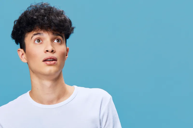 young man looking surprised with curly hair wearing white shirt against blue background, expressing shock and curiosity in bright studio lighting
