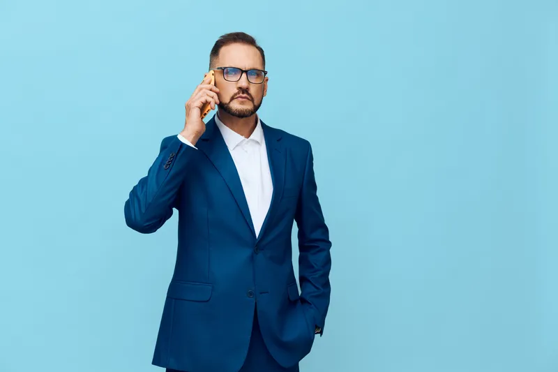 A professional businessman in a navy suit speaks on a mobile phone against a solid blue background, projecting confidence, focus, and calm assurance in a modern corporate setting.