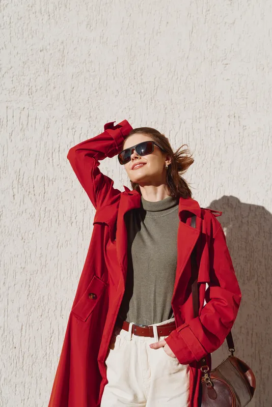 Confident woman outdoors in a red coat, olive turtleneck, white pants, sunglasses, standing relaxed in sunlight against a warm wall