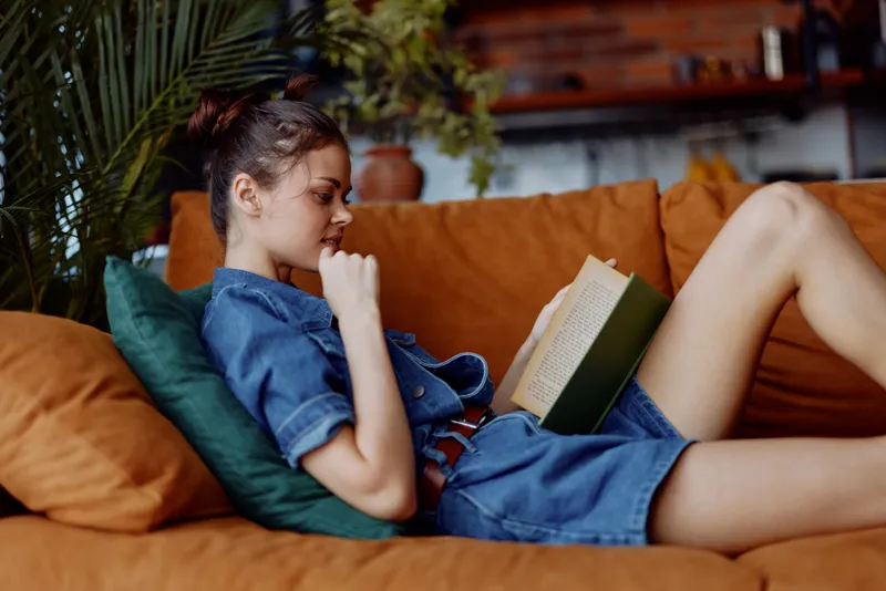Женщина в джинсовой рубашке и очках читает книгу, сидя на диване в уютной домашней обстановке