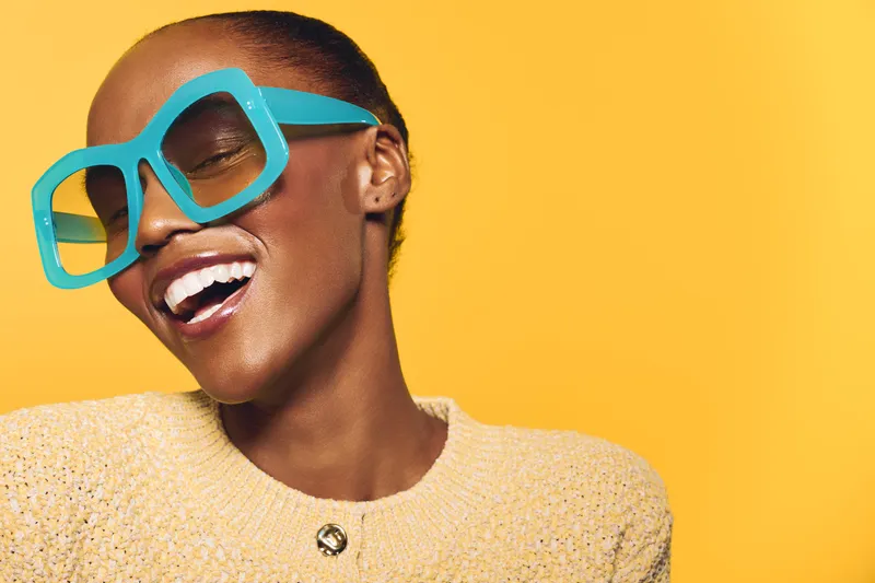 Happy African American woman with dark skin wearing large turquoise glasses smiles with closed eyes on yellow background. Concept for joy, fashion, and beauty in studio lighting.
