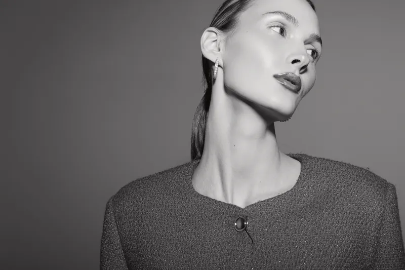 Portrait of a young Caucasian woman with light skin wearing earrings and a textured top, looking sideways on a grey background.