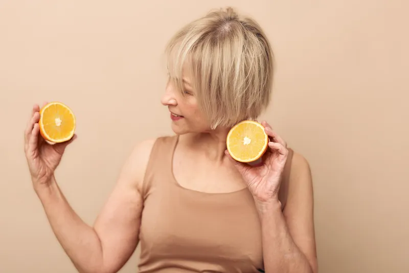 Mature woman with short blonde hair holding two halves of fresh orange fruit smiling and looking at the citrus on beige background. Concept of healthy lifestyle and natural vitamins.