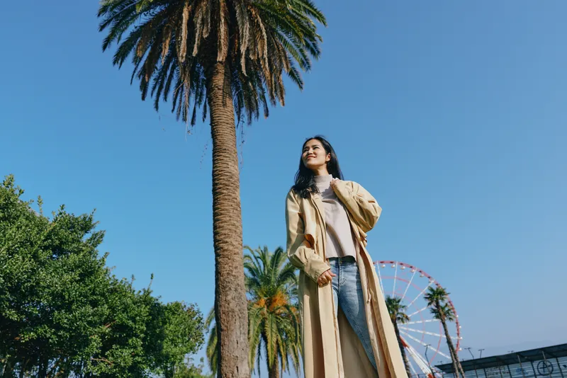 Woman in a long beige coat stands among palm trees under a clear blue sky, outdoor fashion moment with a distant ferris wheel, embodying travel style and sunny leisure