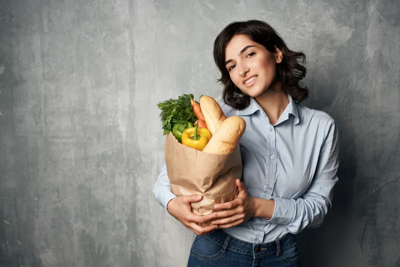 Брюнетка в синей рубашке с пакетом продуктов, в котором лежат овощи, на темном фоне