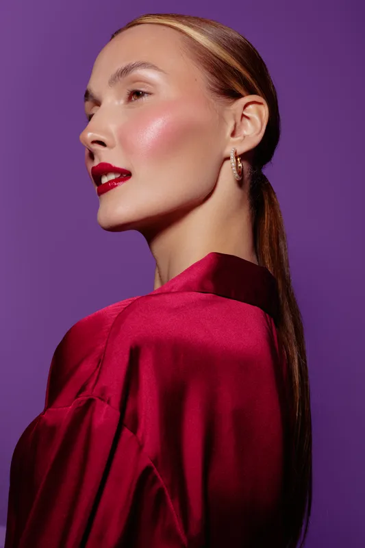 Profile of elegant woman with shiny skin and red lipstick wearing red satin blouse and hoop earrings on purple background. Concept of beauty, fashion, and glamour.