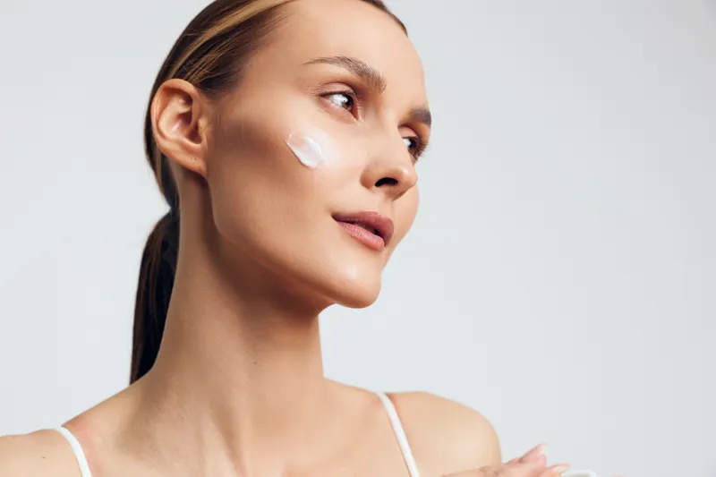 Young woman with clear skin applying white face cream on cheek. Close-up portrait on light background. Concept of skincare, beauty routine, and natural wellness.
