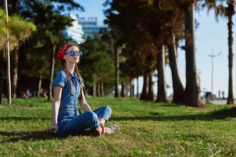 Lifestyle woman in modern boho-western denim street style sitting on grass outdoors, wearing sunglasses and red bandana, enjoying sunny day with film color effect.