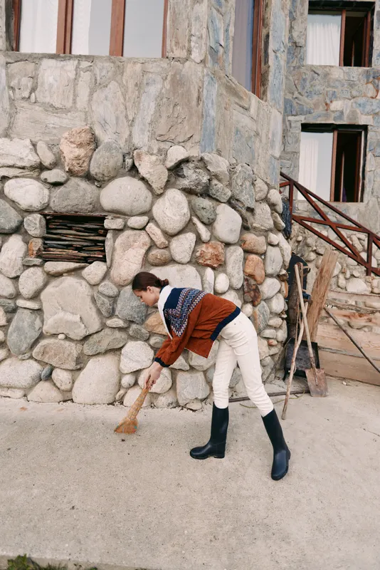 Woman sweeping with broom along stone wall outside a rustic building, wearing sweater and boots while performing outdoor cleaning task on sidewalk in casual autumn scene.