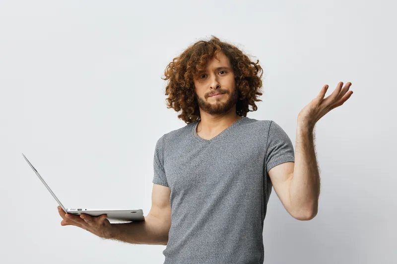 Confused young man with curly hair holding a laptop, expressing uncertainty in a neutral setting, perfect for themes of technology and doubt