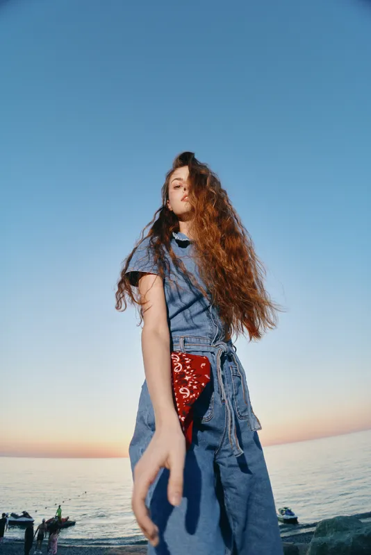 Lifestyle woman in modern boho-western denim street style with long curly hair captured in film color near the ocean during sunset, showcasing casual and natural fashion vibe.
