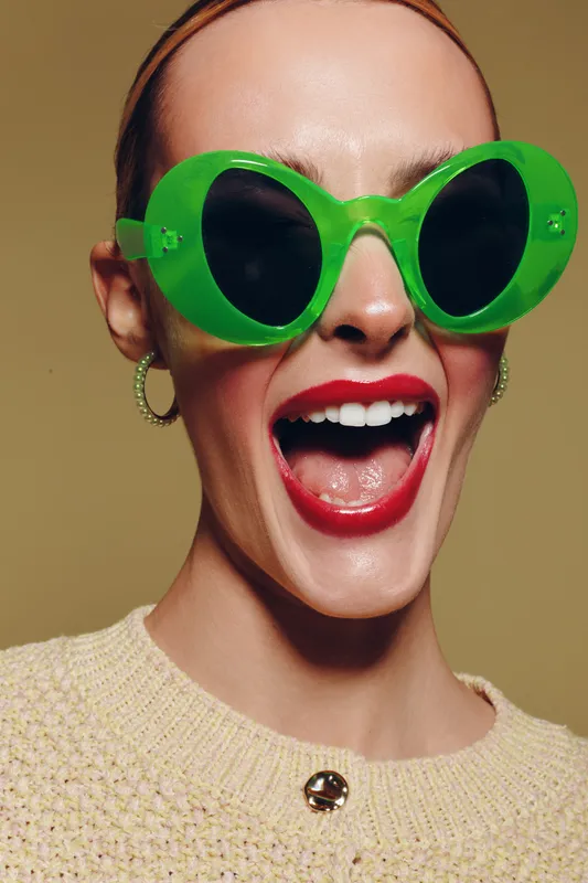 Close-up of a smiling woman wearing large green sunglasses and red lipstick. She wears a beige sweater and hoop earrings against a neutral background. Concept of fashion and joy.