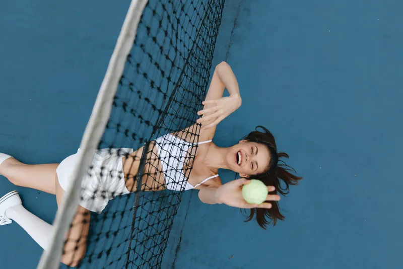 Joyful female tennis player reaching for the ball on a blue court, wearing a stylish athletic outfit and demonstrating enthusiasm and energy in motion