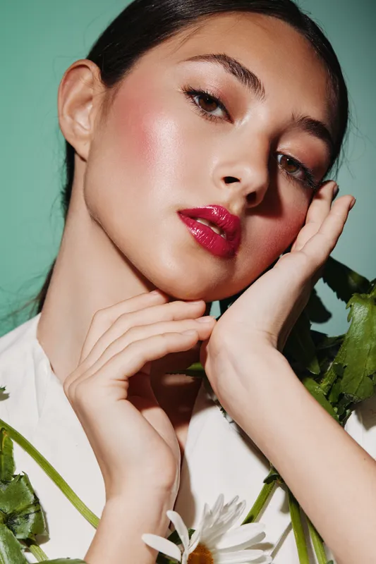Portrait of a fashionable woman with glossy red lips and glowing skin. She poses softly touching her face with hands in studio light. Concept of beauty, fashion, makeup.
