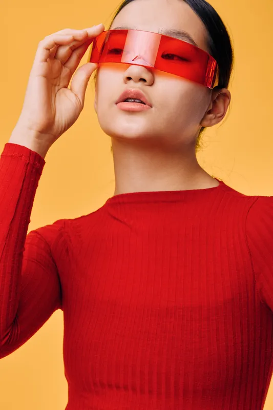 Woman with red transparent glasses and red sweater posing confidently against yellow background. Closeup portrait, fashion style and modern eyewear with hand adjusting sunglasses.