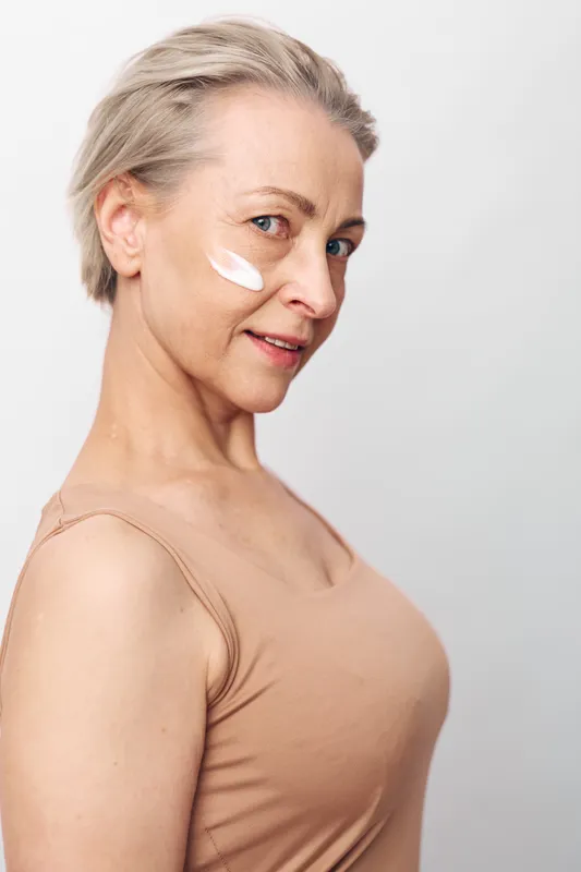 Mature woman with short blonde hair applies white face cream on cheek, wearing beige tank top. Skincare concept focused on natural beauty and anti-aging care with soft studio lighting.