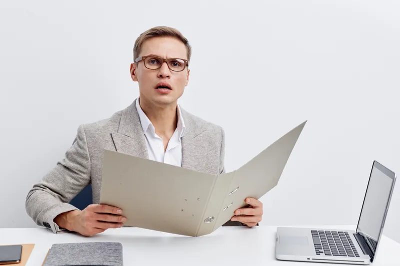 Businessman in formal attire with glasses looking surprised while reading a large document at office desk with laptop, minimalistic background, professional workspace, corporate environment, modern st