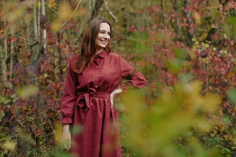 Candid portrait of a smiling woman in a simple red dress standing among autumn woodland foliage, conveying a genuine presence, natural expression and warm authentic connection with the environment.