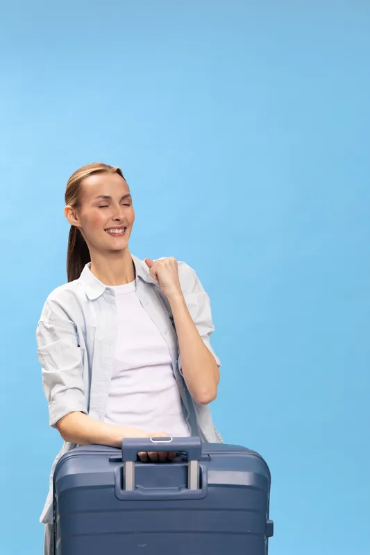 Young woman with ponytail holding blue suitcase smiling with closed eyes on blue background. Concept of travel, vacation, tourism, and summer holiday preparation with relaxed mood.