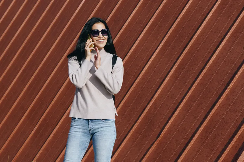 Woman wearing jeans and sweater uses phone, sunglasses, standing against a diagonal wooden wall, urban street style, outdoor lifestyle and casual chic.
