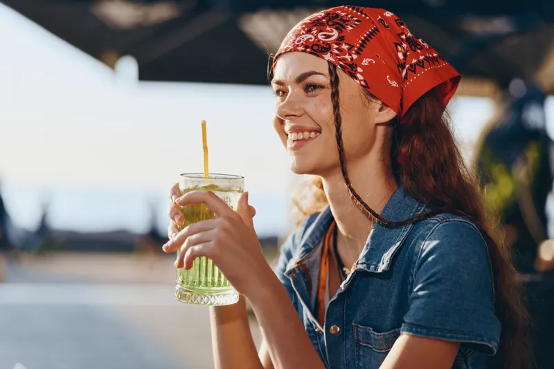 smiling woman with red bandana enjoying a green drink outdoors in warm sunlight, casual denim shirt, relaxed mood, summer atmosphere