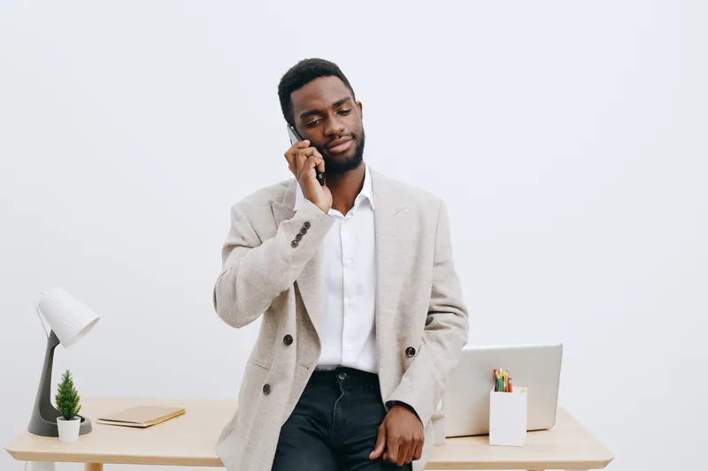 Confident young African American man in beige blazer and white shirt talking on smartphone while standing at modern desk with office supplies isolated on plain white background