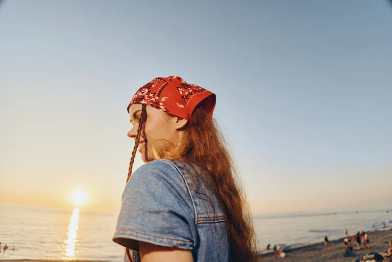 Lifestyle woman in modern boho-western denim street style with red bandana, enjoying sunset at the beach captured in warm film color tones.