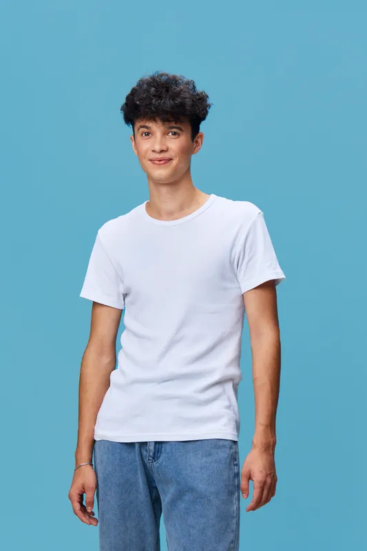 Young man with curly hair wearing a white t-shirt and blue jeans standing against a plain blue background with a relaxed and casual posture.
