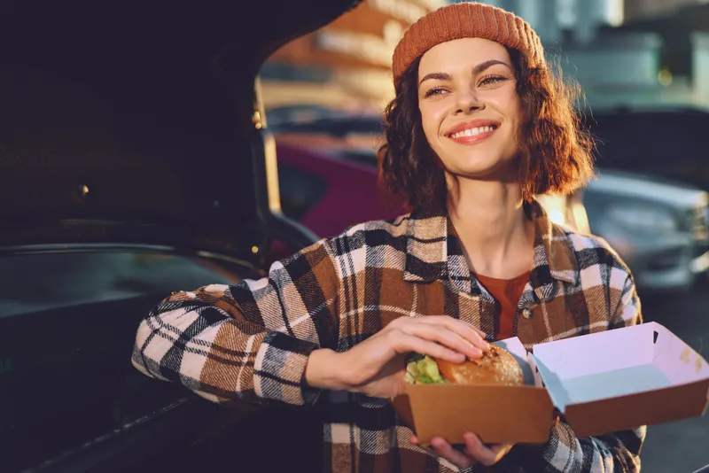 woman smiling with burger takeaway by car in golden hour, beanie and plaid jacket, candid lifestyle portrait with authenticity and emotional storytelling, mindful living urban vibe.