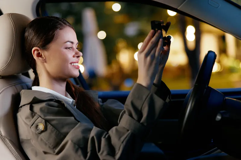 Женщина, автомобиль, мобильный телефон, напиток: женщина сидит на водительском сиденье автомобиля, держа в одной руке мобильный телефон, а в другой — напиток, что отражает момент многозадачности во…