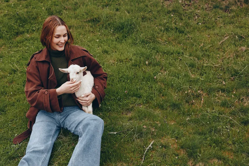 A joyful woman sits on a grassy hillside, holding a kid goat close in her arms, smiling softly. A relaxed, rural lifestyle moment highlighting harmony between people and animals outdoors.