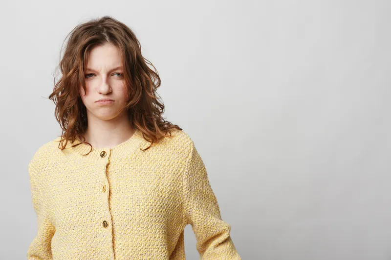 Young woman with curly hair wearing yellow sweater looks frustrated and angry isolated on grey background. Concept of negative emotions, stress, and dissatisfaction.