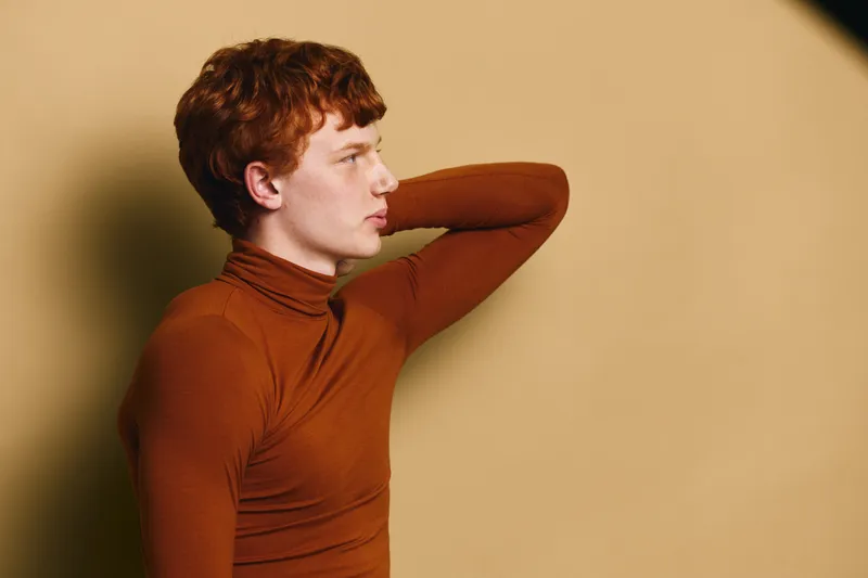 Young man with red hair and pale skin wearing brown turtleneck looking side in studio with beige background. Casual pose with hand behind head and thoughtful expression.