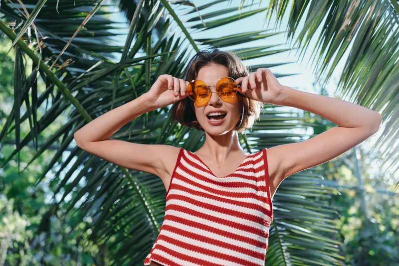 A cheerful woman wearing a striped red and white tanktop stands outdoors among palm leaves, adjusting round orange sunglasses with a playful smile, exuding sunny summer energy.