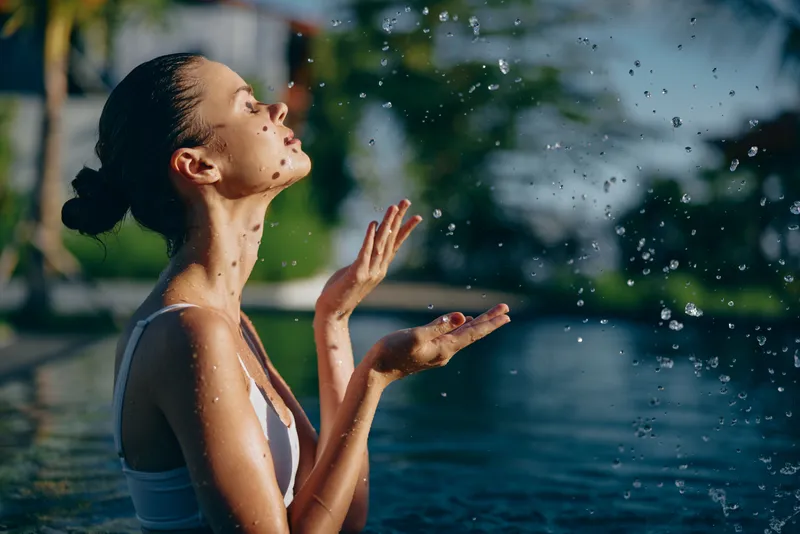 Lifestyle shot of a sunlit woman by a pool, relaxing as water droplets spray, wearing a pale swimsuit, hair in a bun, eyes closed, enjoying a calm summer breeze and tranquil moment