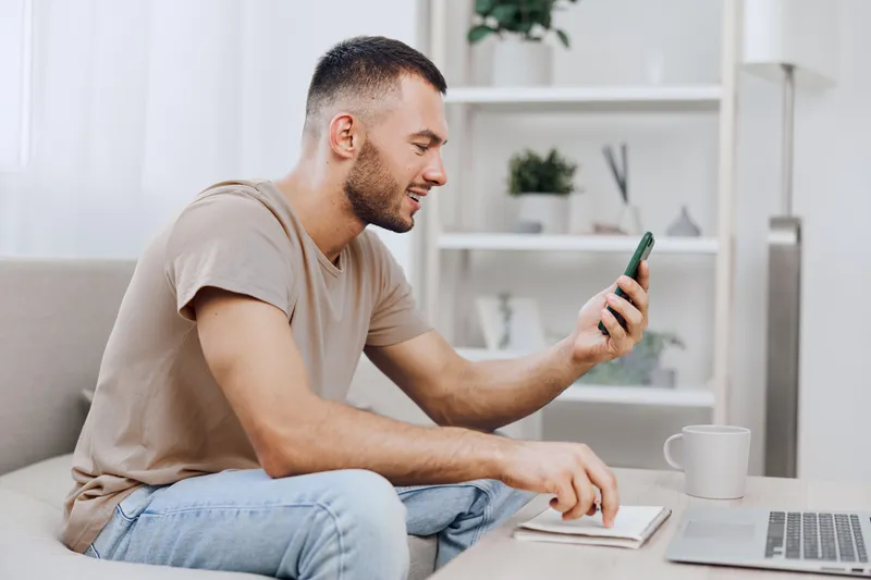 Smiling man using a smartphone while sitting on a cozy sofa, creating a relaxed atmosphere with soft natural lighting and a minimalistic background.