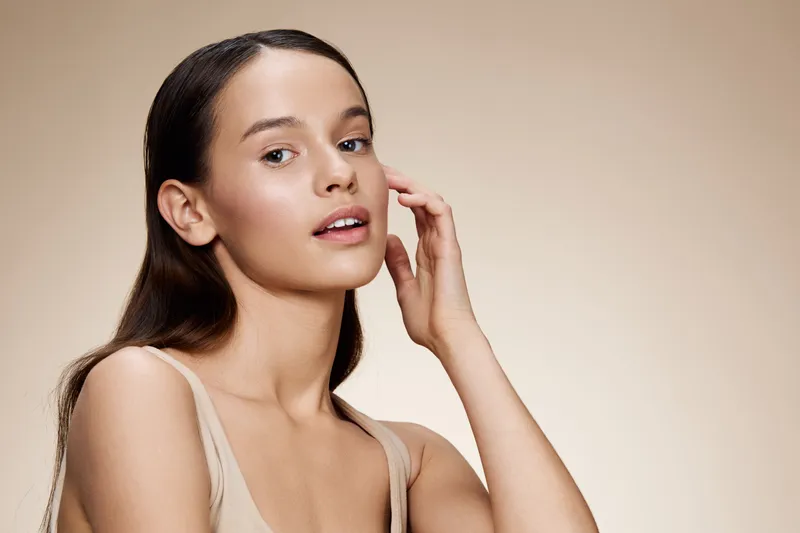 Young woman with natural makeup, wearing a neutral outfit, showcasing healthy skin and a gentle expression against a soft beige background