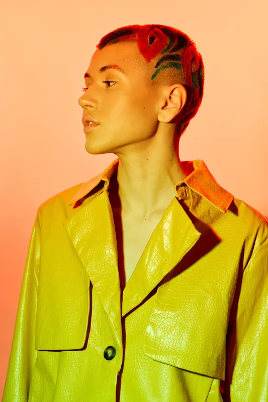 Woman with creative red hairstyle and short hair wearing a yellow shiny jacket, posing confidently against a warm orange background with dramatic side lighting.