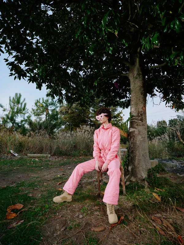 Outdoor portrait of a woman in a pink jumpsuit sitting beneath a large tree in a natural setting, relaxed pose, soft daylight, casual fashion, calm mood, simple composition for lifestyle photography