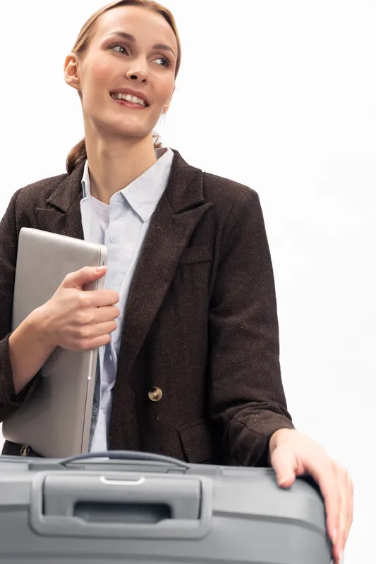 Businesswoman in formal suit holds laptop and suitcase, smiling and looking away. Concept of travel, work, business trip, and professional lifestyle with white background.