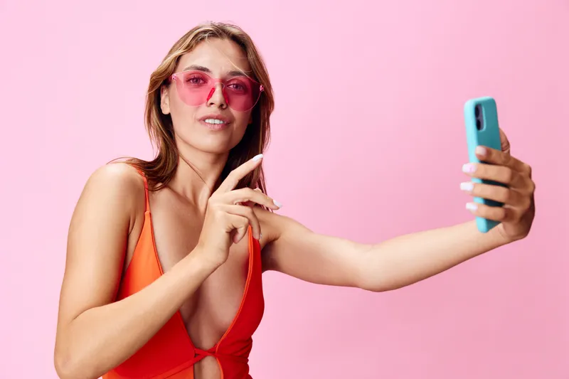Привлекательная молодая женщина в купальнике делает селфи на смартфон на ярком розовом фоне