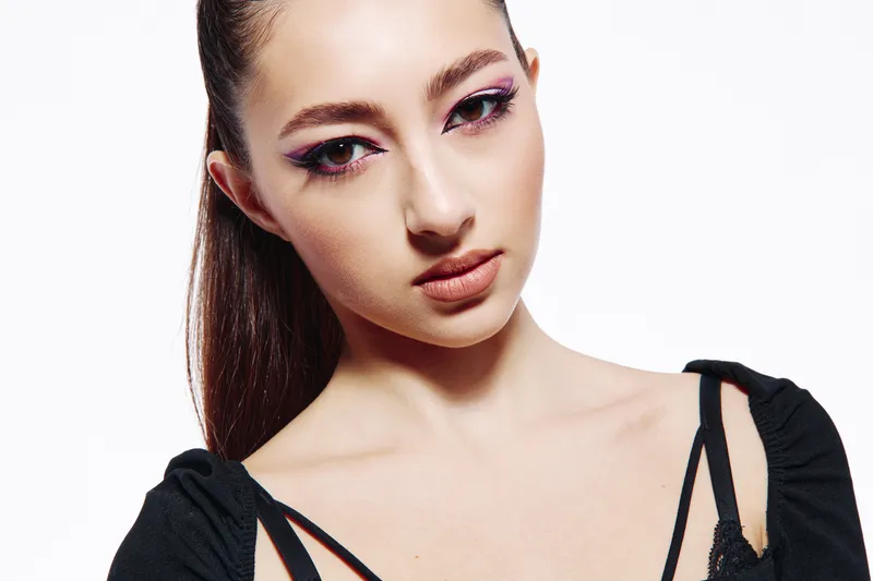 Portrait of a young woman with creative pink eye makeup wearing a fashionable black top. White background. Concept of beauty, fashion, and creative expression.