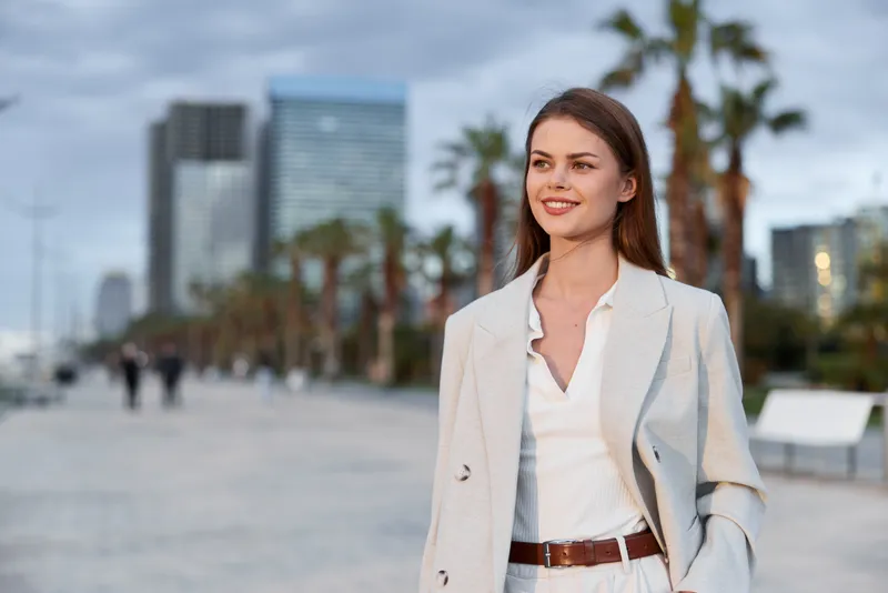 Confident businesswoman smiling outdoors in a modern urban environment, dressed in a stylish light suit and enjoying a sunny day.