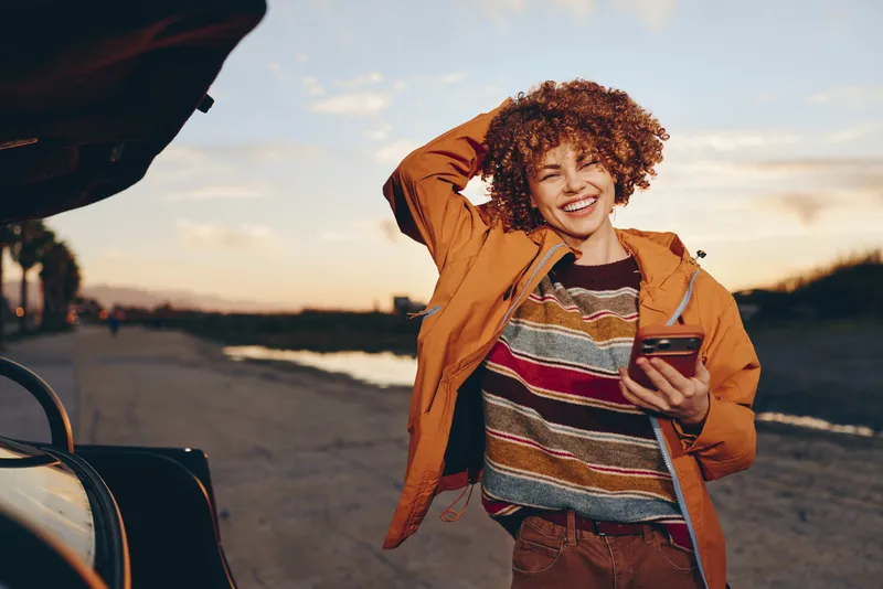 Woman outdoors, smiling in sunset light, wearing jacket, holding smartphone, cheerful lifestyle near a car during roadtrip, warm golden sky, casual pose open road background, soft wind