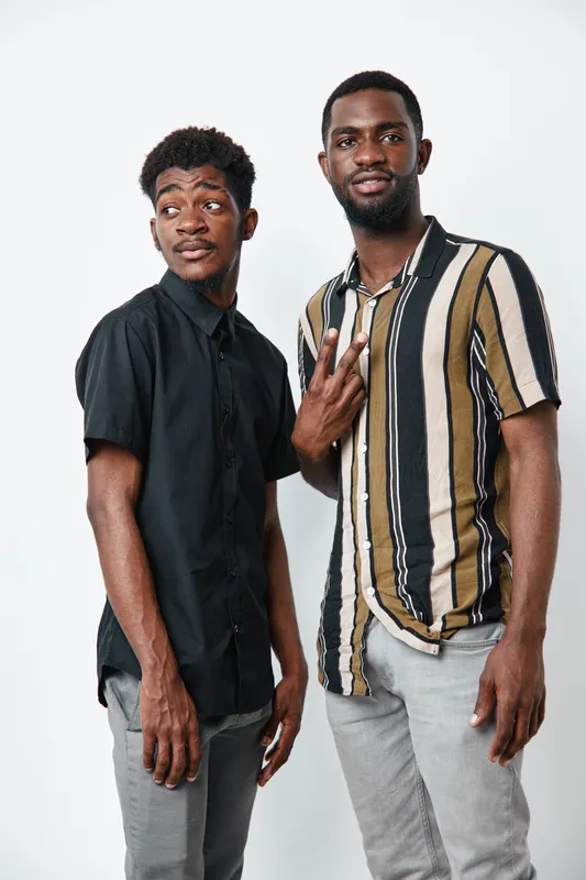 Two African American men with dark skin posing together in casual shirts and gray pants against white background. One man shows peace sign while the other looks sideways.