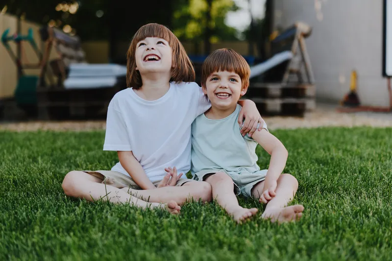 Two joyful boys sitting together on the grass, enjoying their carefree summer day with bright smiles and playful expressions, radiating happiness and friendship.