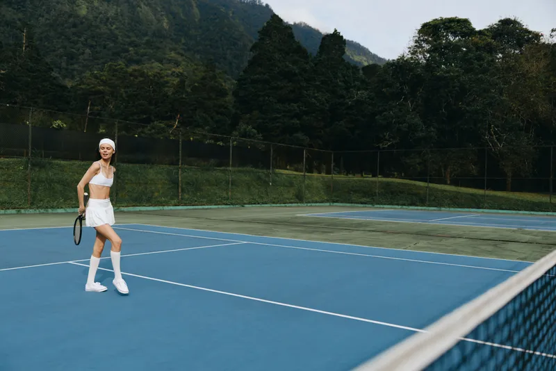 Young athletic woman playing tennis on a blue court, wearing a white sports outfit and headband, smiling with a beautiful green background