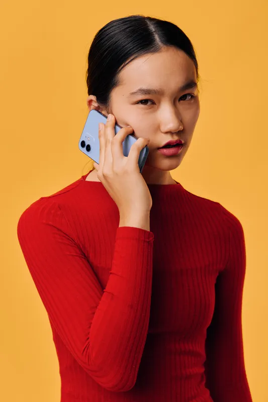 Woman with smartphone in hand talking on phone against orange background. Young female wearing red sweater using mobile device for communication and conversation.