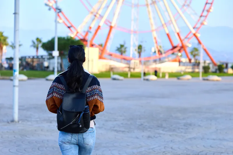 Woman with backpack walks toward a large ferris wheel at an outdoor amusement park, palm trees in distance, casual jacket, travel mood, exploration vibe, open space, leisure scene, bright daylight