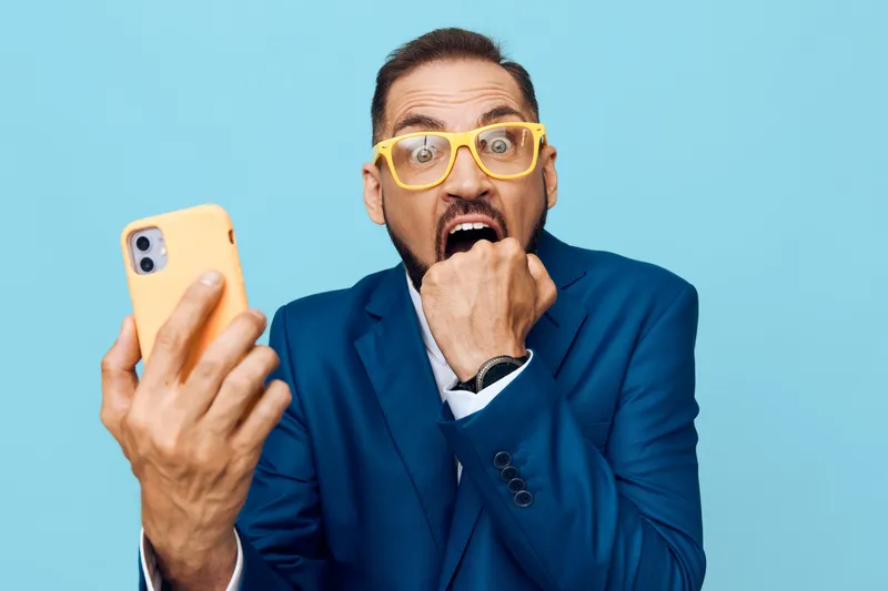 A professional businessman in a blue suit stands against a solid colored backdrop, an isolated setting, showing surprise and urgency as he checks his phone.